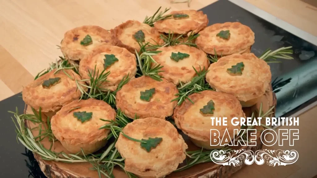 A collection of golden-brown pastries is displayed on a dark plate, each topped with a small green leafy garnish. Sprigs of rosemary are scattered amongst the pastries. The background shows a portion of a wooden countertop. The logo for 'The Great British Bake Off' appears prominently at the bottom of the image.