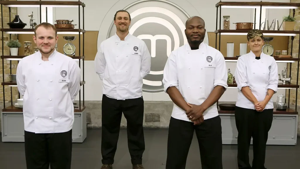 Four contestants from "MasterChef UK" stand in a kitchen setting with decorative shelving behind them. From left to right: A young man with short hair, wearing a chef's jacket with a name tag reading "ELIAN"; a taller man with long hair tied back and a beard, wearing a chef's jacket with a name tag reading "JAMES"; a bald man with a chef's jacket and a name tag reading "OLA"; and a woman with short, wavy hair, wearing a chef's jacket. All are dressed in white chef's jackets and stand confidently in front of the iconic "MasterChef" logo.
