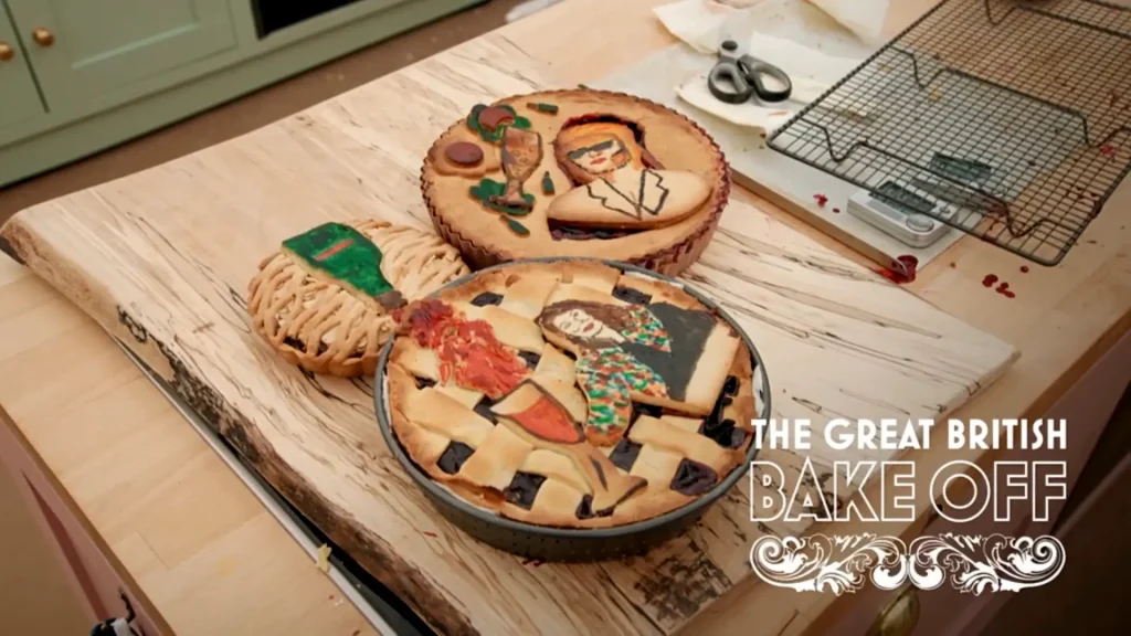 A wooden countertop displays three intricately designed pies. The first pie on the left has a woven top with a green book motif in the center. The middle pie showcases an artistic representation of a person in sunglasses holding a wine glass, surrounded by various objects like a plant. The third pie on the right depicts a detailed scene with a person wearing a hat and colorful attire, surrounded by cut-out shapes. In the background, there are kitchen utensils like scissors and a cooling rack. The logo for 'The Great British Bake Off' is positioned at the bottom right corner of the image.