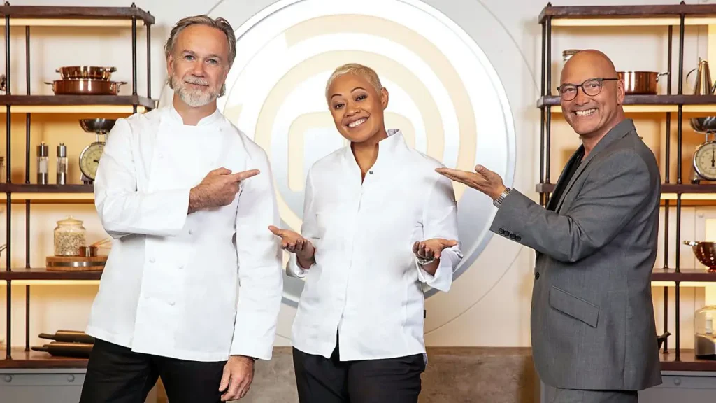 Three individuals from "MasterChef UK" stand in a well-decorated room. The man on the left, with a beard and gray hair, wears a white chef's jacket. The woman in the center has short blonde hair and also dons a chef's jacket. The man on the right is bald with glasses, wearing a gray blazer. All three are smiling and pointing towards the center.