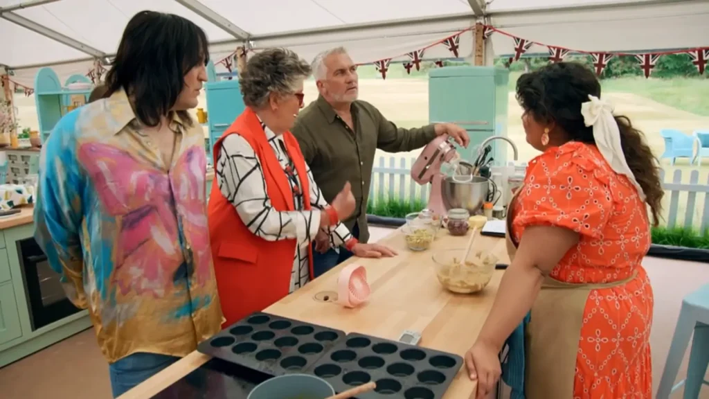 Four individuals inside a baking tent with colorful bunting. From left to right: Noel Fielding with long black hair wearing a pastel-colored shirt with an abstract butterfly design, Prue Leith with short gray hair in an orange jacket and black-and-white patterned shirt, Paul Hollywood in a green shirt holding a pink handheld mixer, and a woman with long dark hair tied with a white ribbon, wearing an orange dress with circular patterns. In front of them is a wooden table with baking tools, a muffin tin, and a bowl of ingredients.