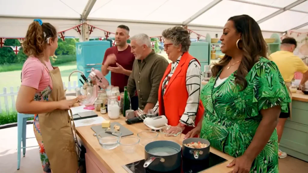 Five individuals in a baking tent adorned with British flags. From left to right: A woman with curly hair tied with a blue ribbon, wearing a pink shirt and an apron with floral accents, stands at a counter with a pink mixer and various baking ingredients. Next, a man in a maroon shirt converses with Paul Hollywood, who is wearing a green shirt and visibly discussing something about the baking process. Adjacently, Prue Leith, with gray curly hair, stands wearing an orange jacket and a striped shirt, while handling a white mixer. On the far right, Alison Hammond in a vibrant green dress with a tropical leaf pattern is captured mid-conversation. In the background, another person in a yellow shirt can be seen at another baking station.
