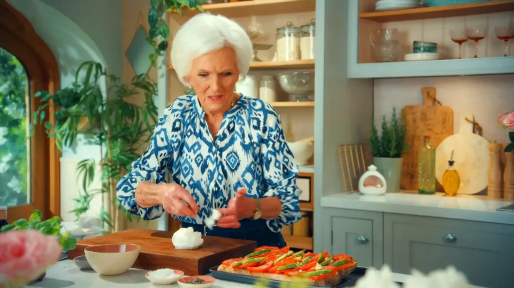 In "Mary Makes It Easy Episode 1 - Weekend Wonders," Mary, an elegant older woman with striking white hair, is meticulously crafting a dish in her sunlit, well-appointed kitchen. She's dressed in a vibrant blue and white patterned blouse, and as she attentively places cream on a dish, her surroundings reveal a blend of rustic and modern kitchen decor. The shelves behind her hold neatly organized kitchenware, while green indoor plants by the window add a touch of freshness. The ambiance is one of warmth, expertise, and the joy of home-cooked meals.