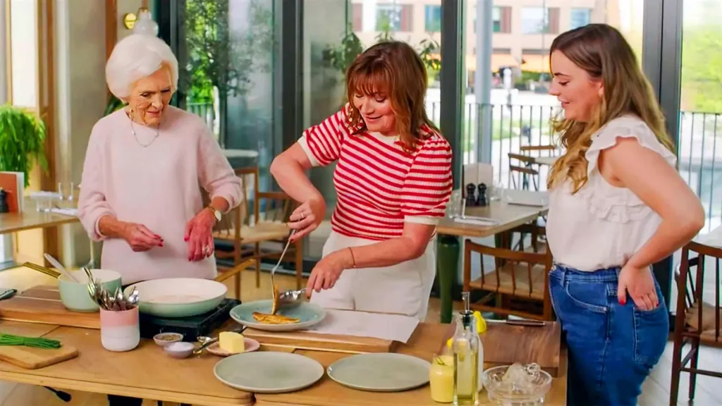 Three women of different generations cooking together in a bright kitchen. The eldest, possibly a cooking show host, oversees the middle-aged woman frying something in a pan, while a younger woman watches with a smile. The kitchen is modern and well-lit, with fresh ingredients and cooking utensils neatly arranged on the counter.