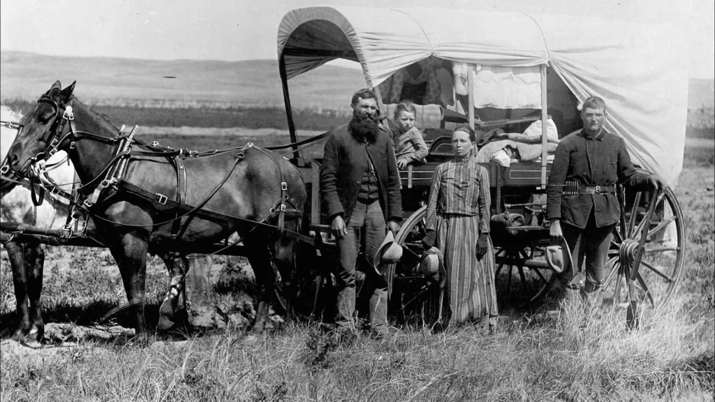 A black and white historical photograph showing a pioneer family standing beside their horse-drawn covered wagon on open grassland. The image features a horse in harness at the left, three adults and what appears to be a child standing beside the wagon. The wagon has a distinctive white canvas cover stretched over hoops. The terrain is grassy with rolling plains visible in the background, typical of the American frontier landscape.