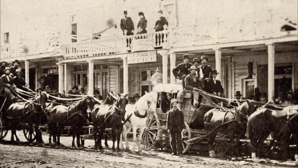 A historical black and white photograph showing multiple horse-drawn stagecoaches lined up in front of a Western frontier town building. The wooden structure features a visible 'STAGE CO'S OFFICE' sign and a second-story balcony where three individuals stand. Several men in period attire including formal suits and hats are positioned on and around the stagecoaches. Multiple horses, including a distinctive white horse in the center, are harnessed to the coaches. The dirt road in front and wooden storefronts with columns create a classic scene of transportation in the American frontier era.